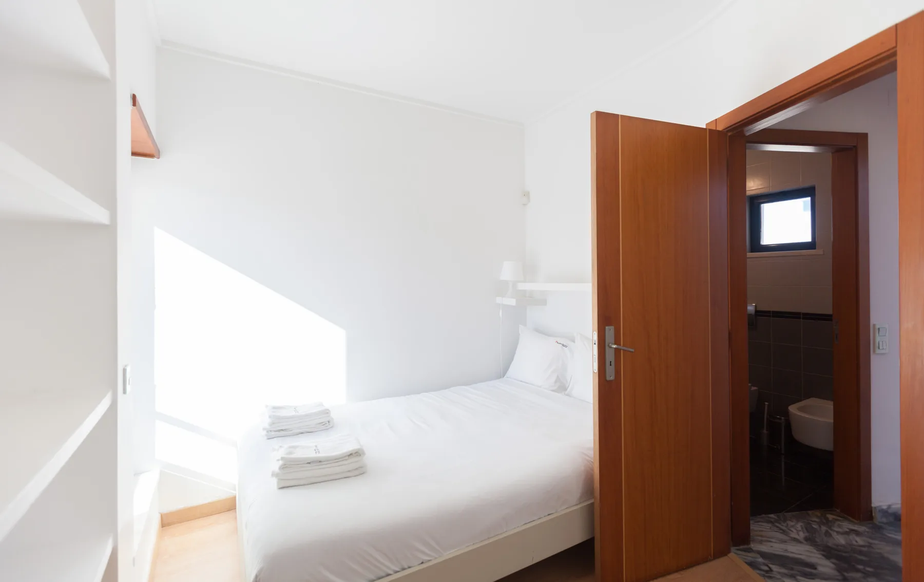 A bright bedroom with white bedding and an open door to a bathroom.