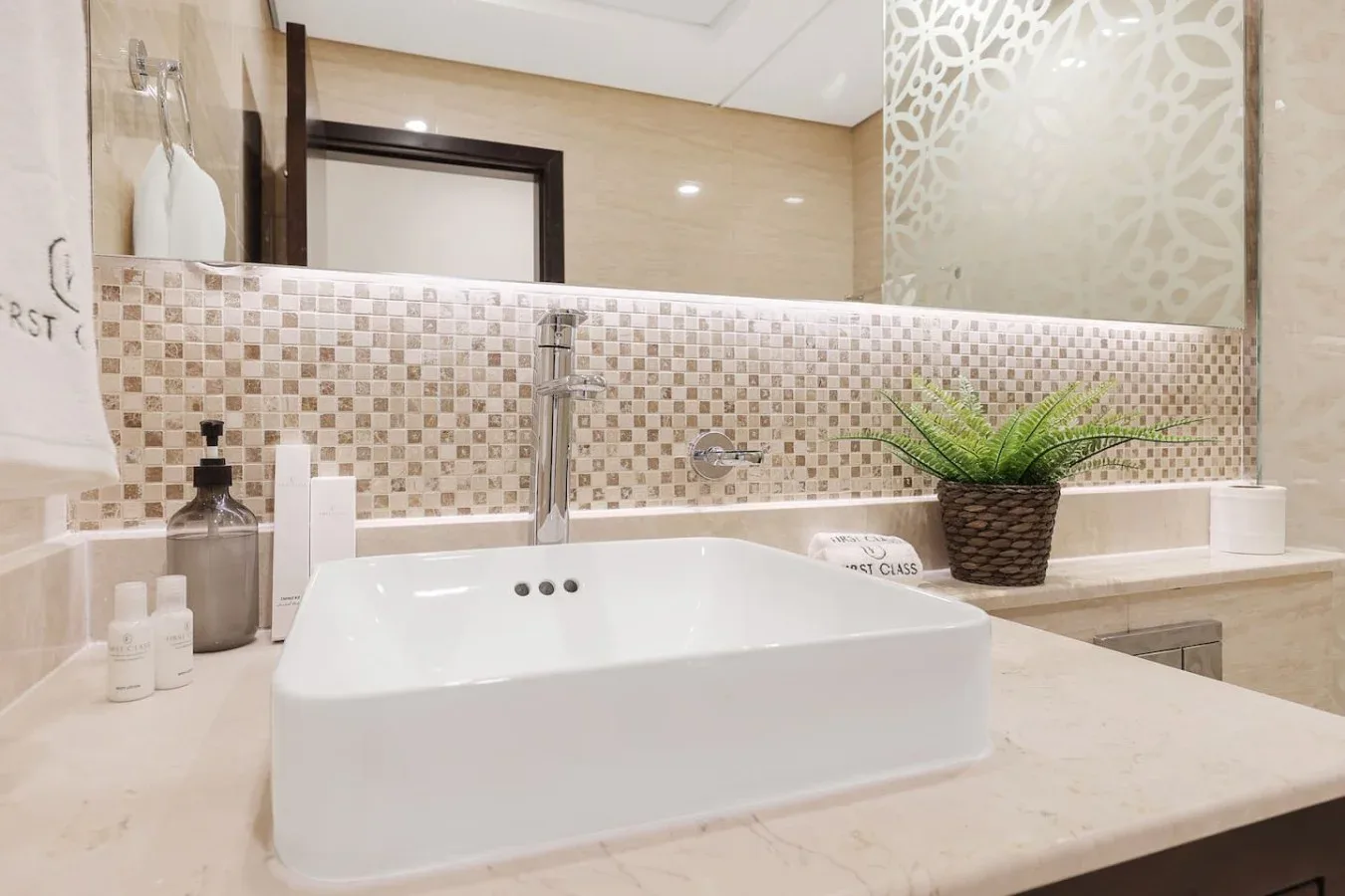 Un tocador de baño con lavabo blanco sobre encimera, pared de azulejos y una planta.
