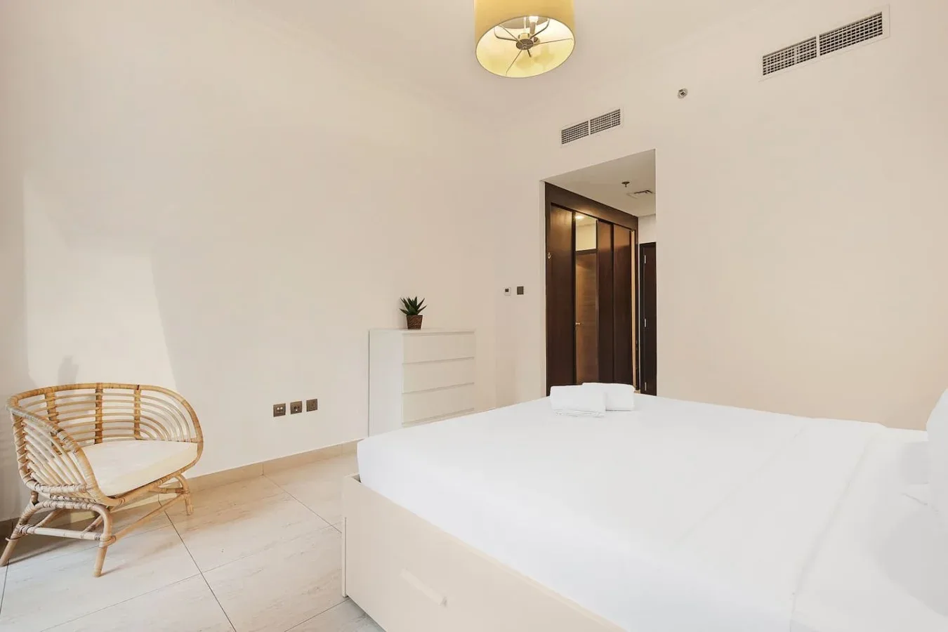 Un dormitorio luminoso con una cama blanca, una silla de mimbre y una cómoda pequeña.