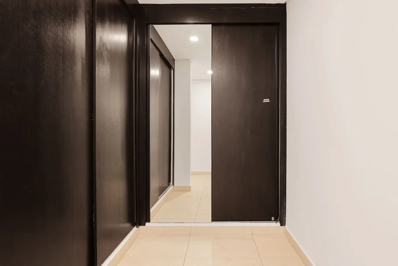 Un pasillo con puertas oscuras da a un corredor más luminoso con suelo de baldosas.