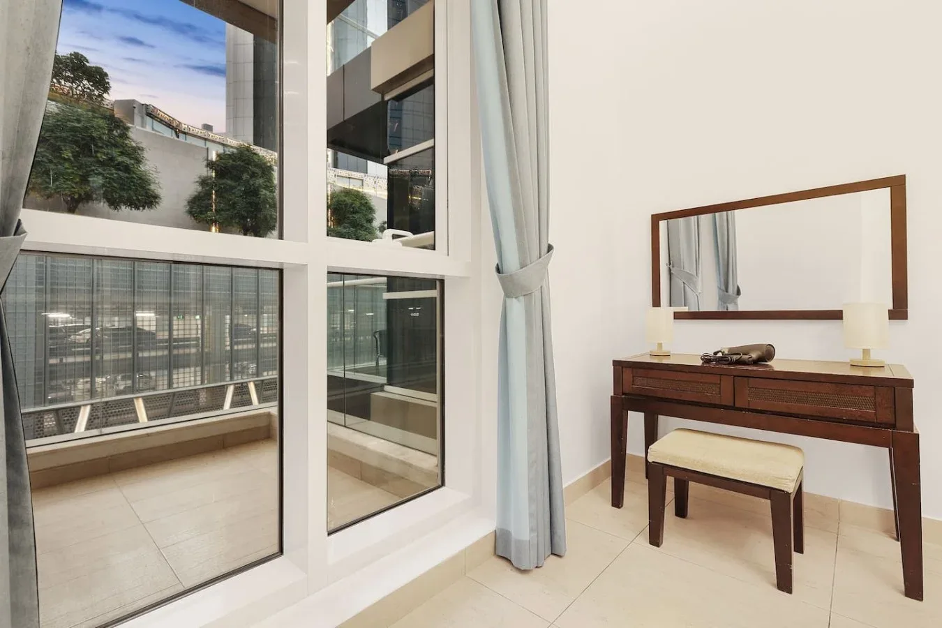 Un dormitorio luminoso tiene un tocador de madera y una ventana grande con vistas a edificios cercanos.