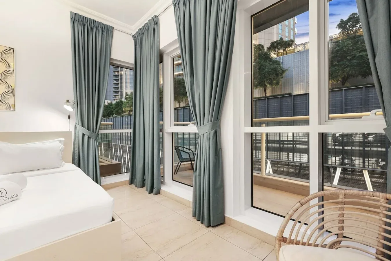 Un dormitorio luminoso con ventanas altas y cortinas que dan a edificios cercanos.