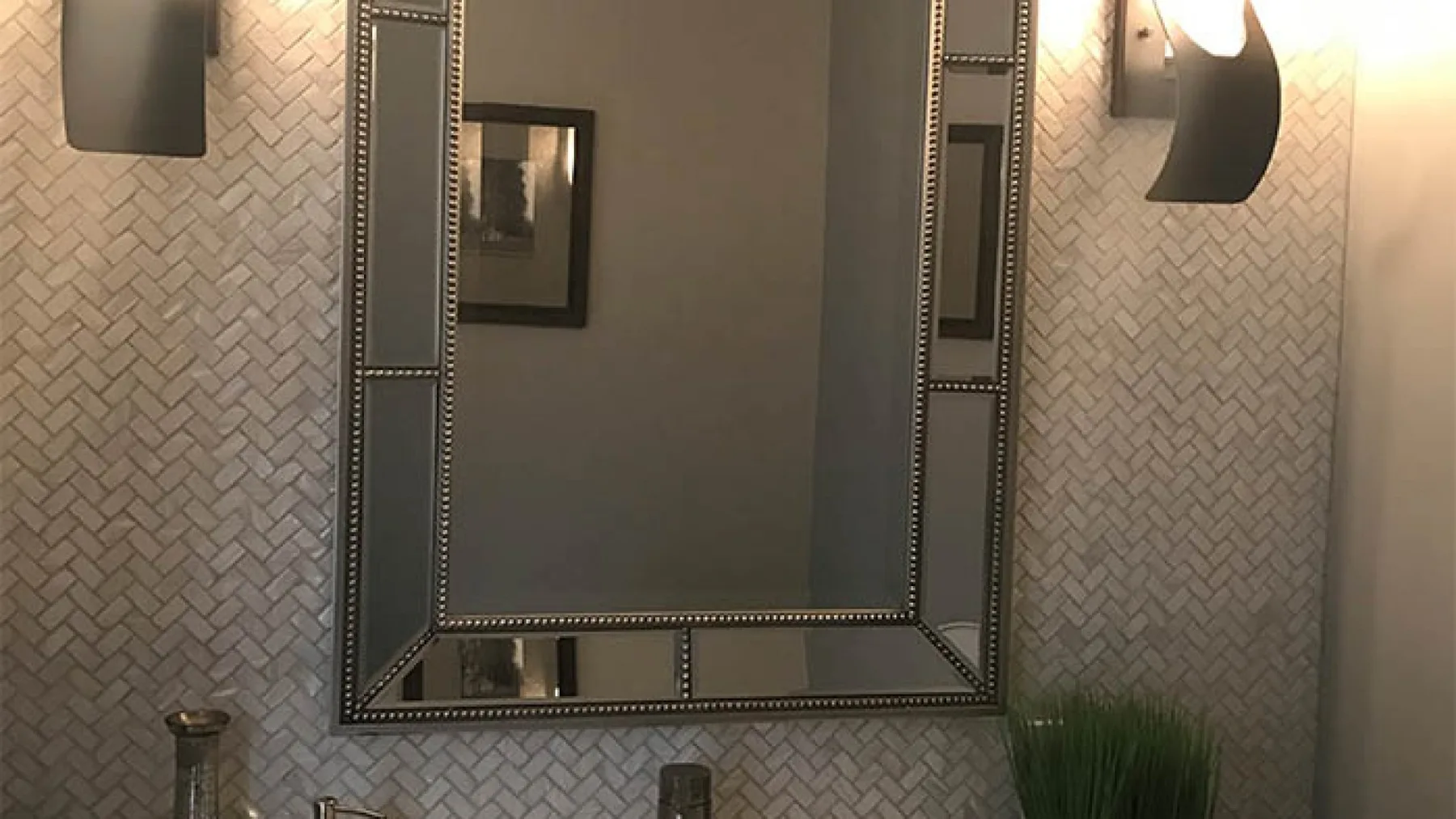 A bathroom mirror hangs above a tiled wall beneath wall lights.