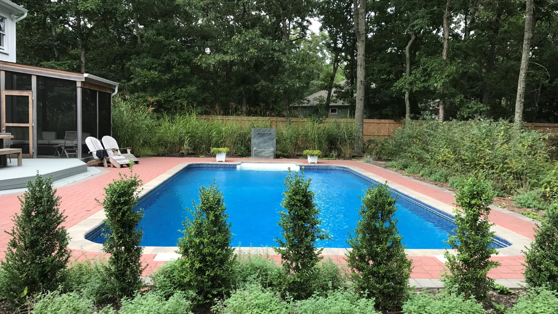 A backyard swimming pool is surrounded by a paved deck, lounge chairs, and greenery.