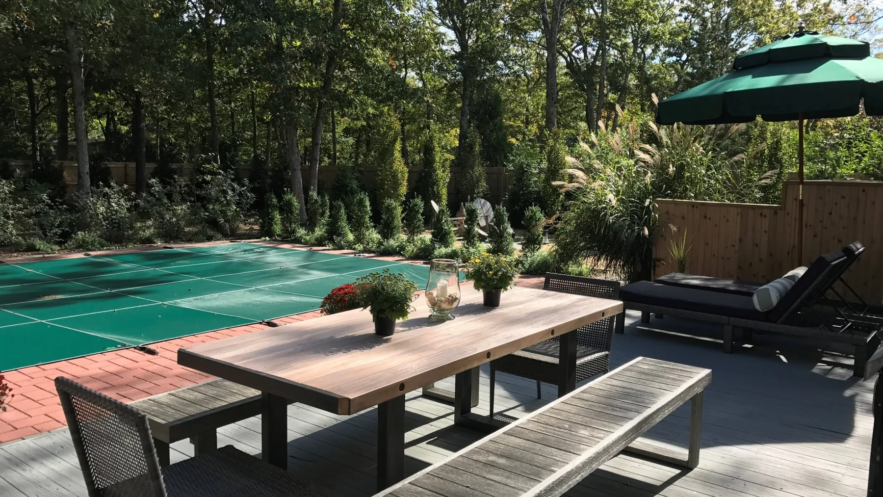 A shaded outdoor deck has a long table, benches, and a green court.