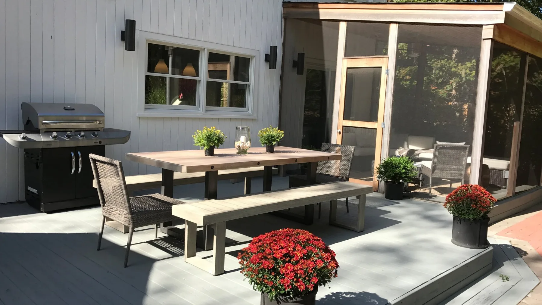 An outdoor patio has a dining table, grill, and potted flowers beside a glass-enclosed area.
