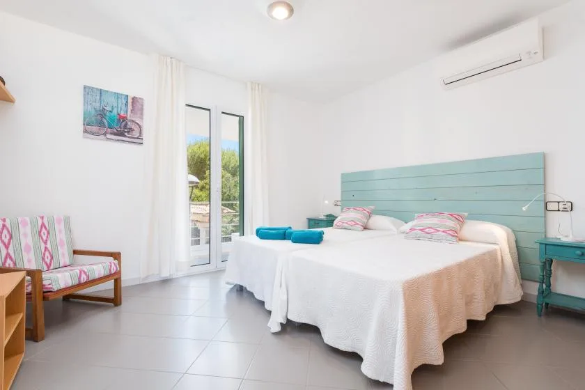 A bright bedroom with two beds, a turquoise headboard, and a window.