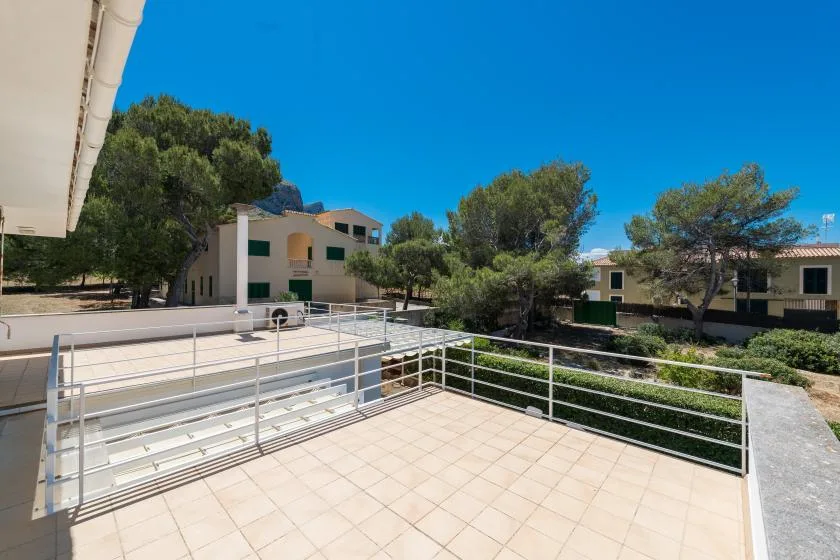 An outdoor tiled terrace overlooks low buildings and trees beneath a clear blue sky.