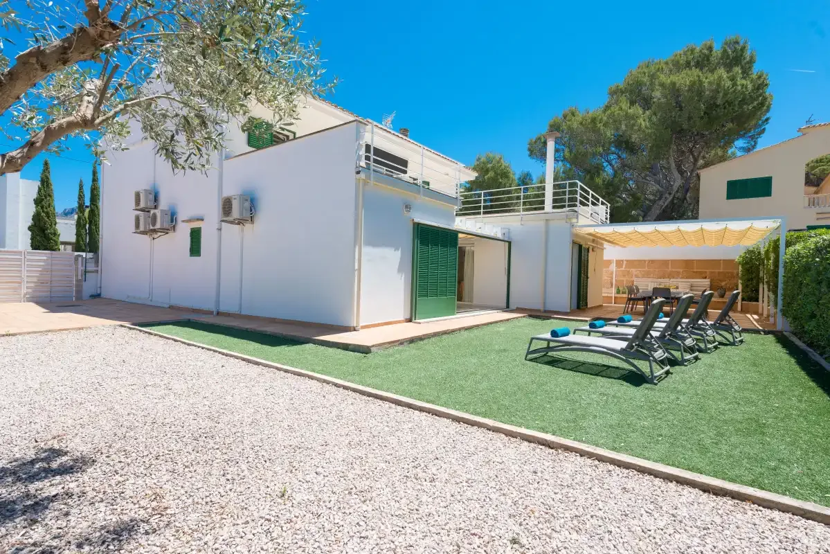 A modern white house opens onto a lawn with sun loungers and a shaded patio.