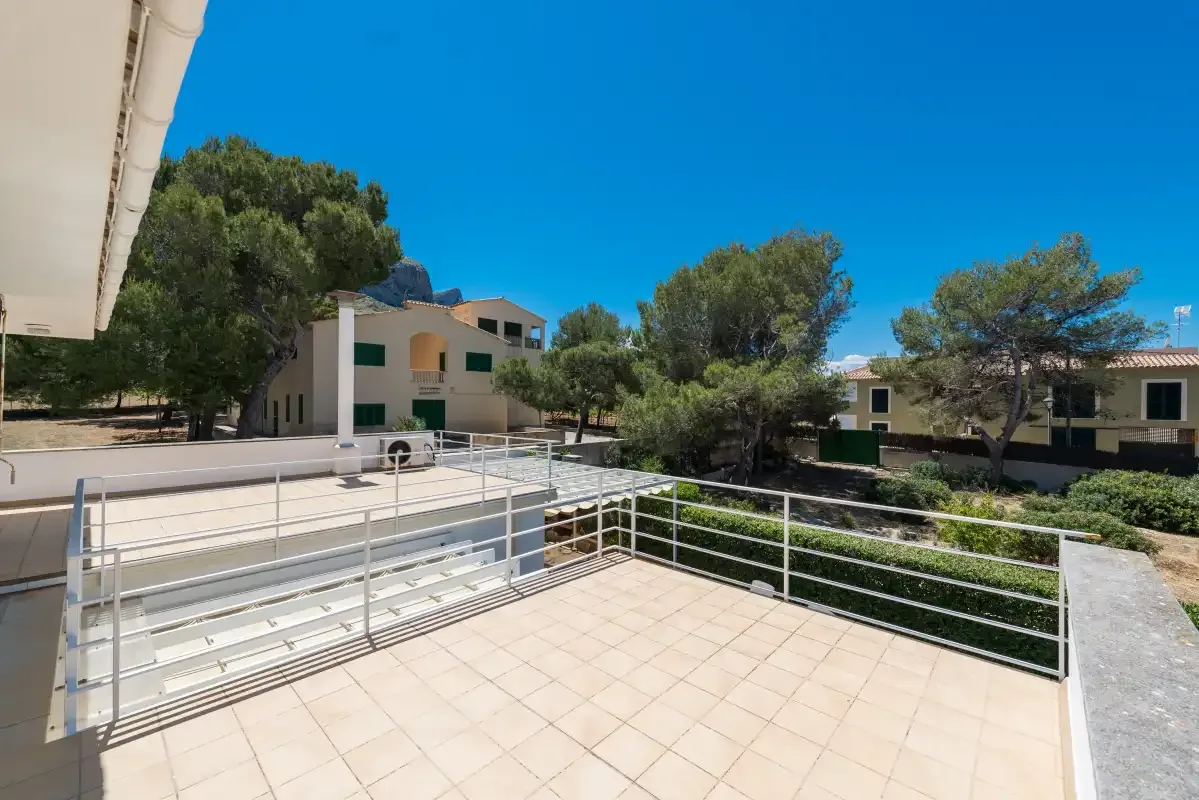 A tiled outdoor terrace overlooks landscaped grounds and low buildings beneath a clear blue sky.