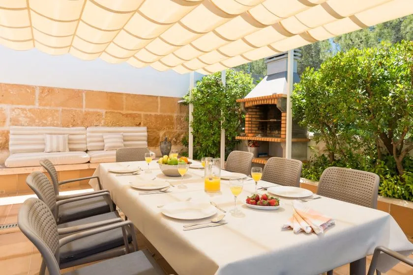 An outdoor dining area has a long set table beneath a canopy, with greenery nearby.