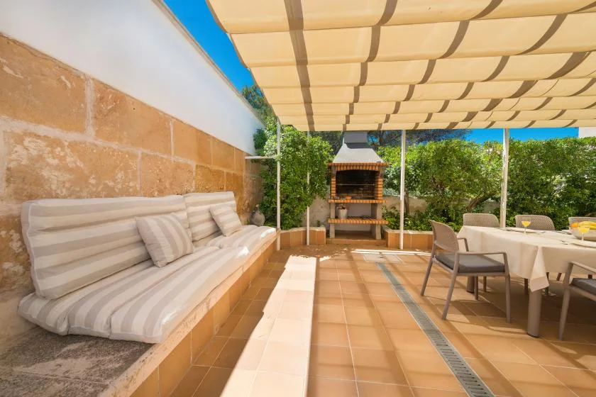 A covered outdoor patio with cushioned seating, a dining table, and a brick barbecue.