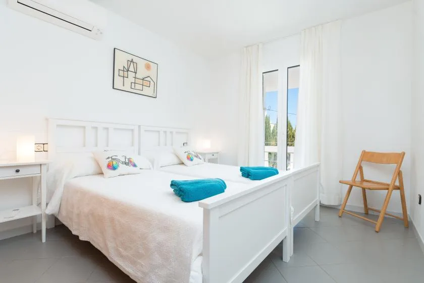 A bright bedroom with white bedding, a tall window, and a wooden chair.