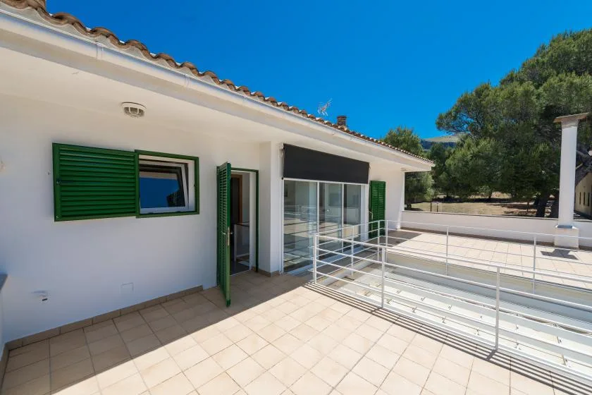 A tiled outdoor terrace beside a white building, with green shutters and a glass doorway.
