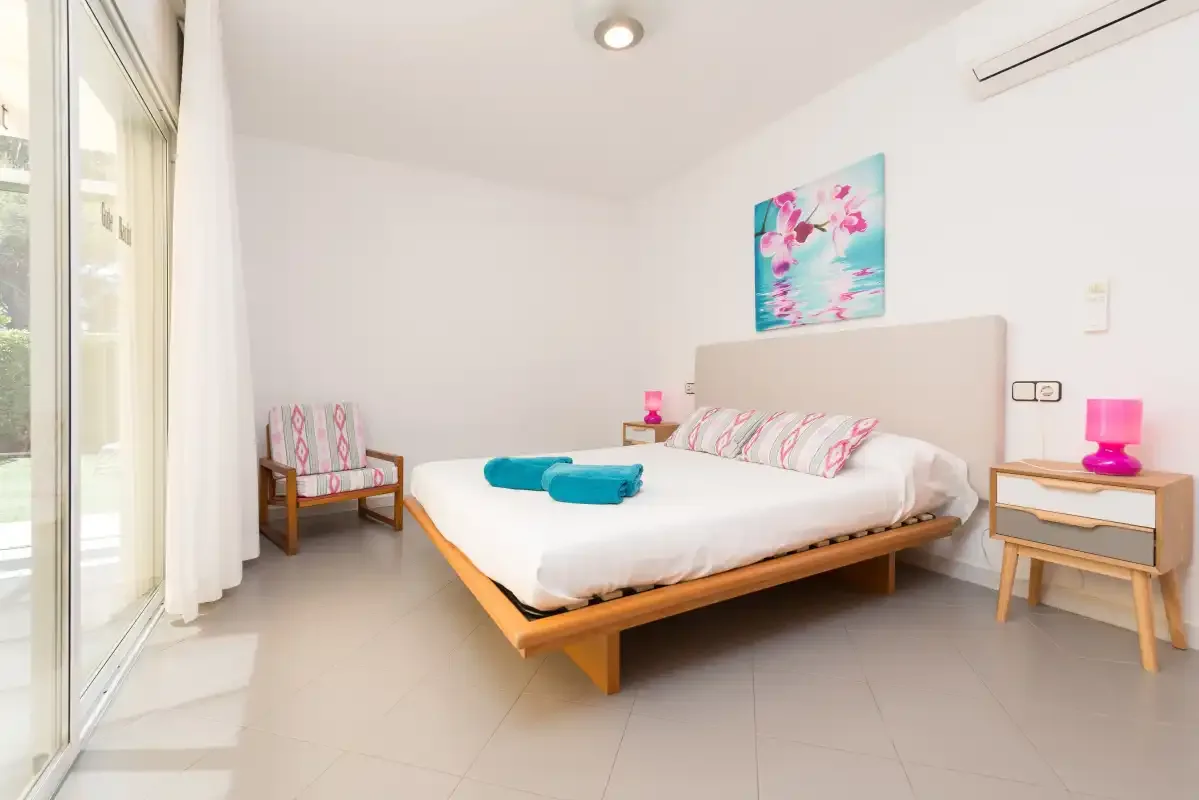 A bright bedroom with a bed, colorful artwork, and a chair beside the window.
