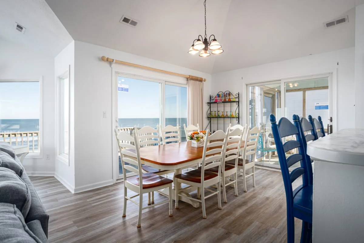 Un comedor luminoso tiene una mesa larga y sillas junto a ventanas con vistas al agua.