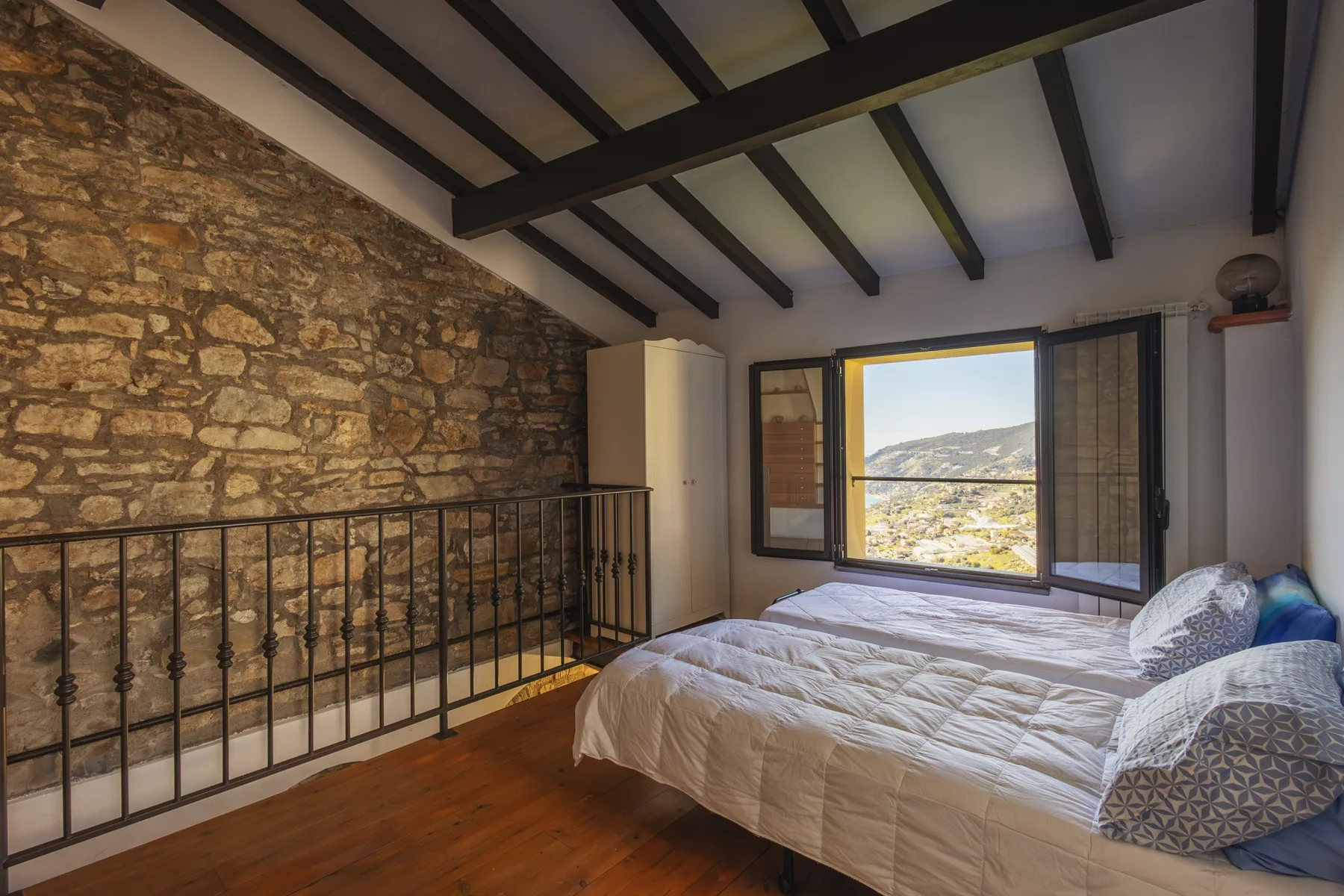 A bedroom with a bed beside a stone wall and an open window overlooking the landscape.