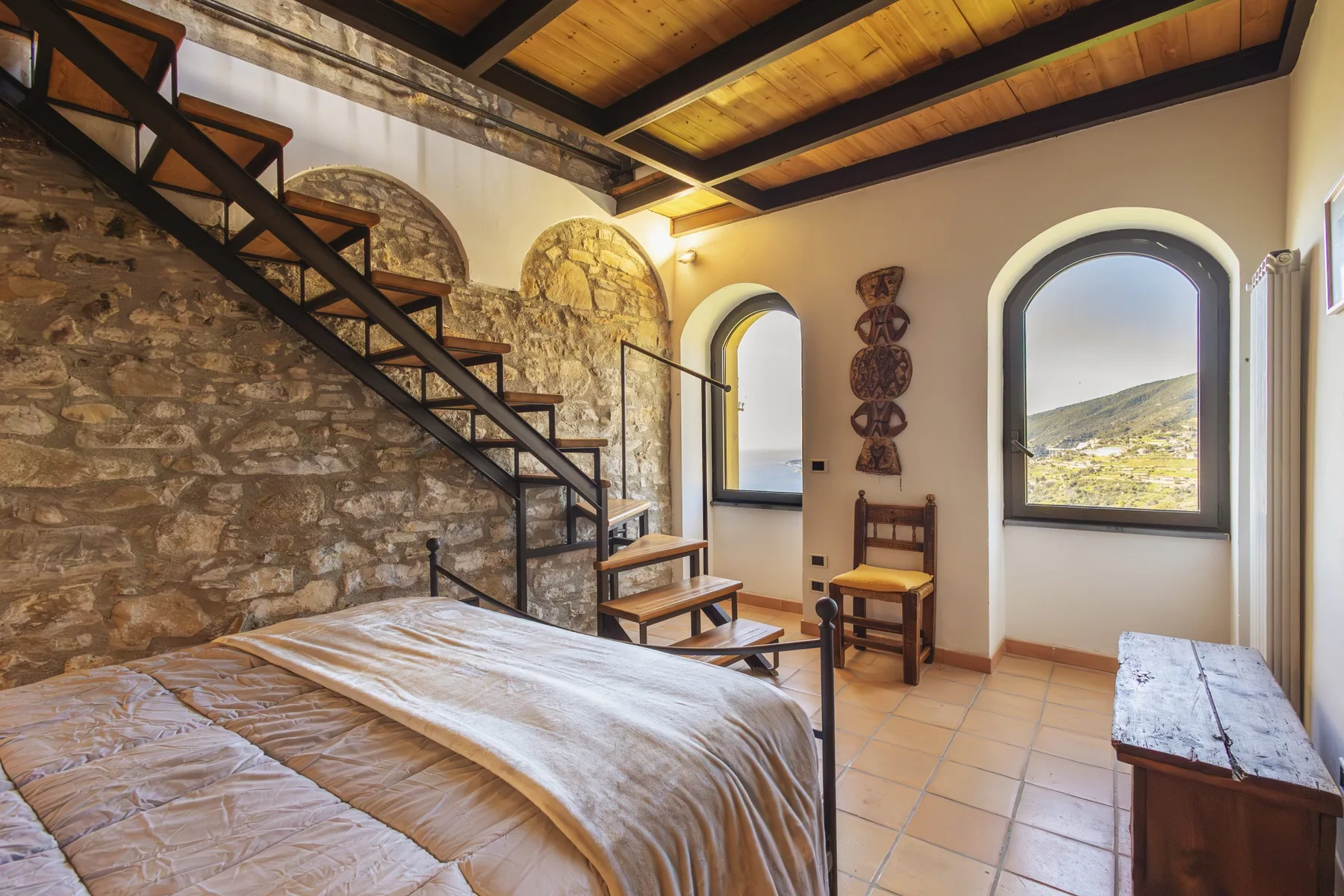 A bedroom with a bed, stone wall, and dark wooden ceiling beams.