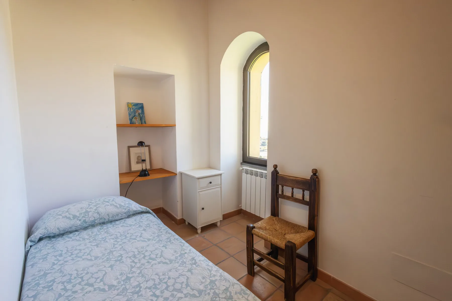 A bedroom with a bed, a wooden chair, and an arched window.