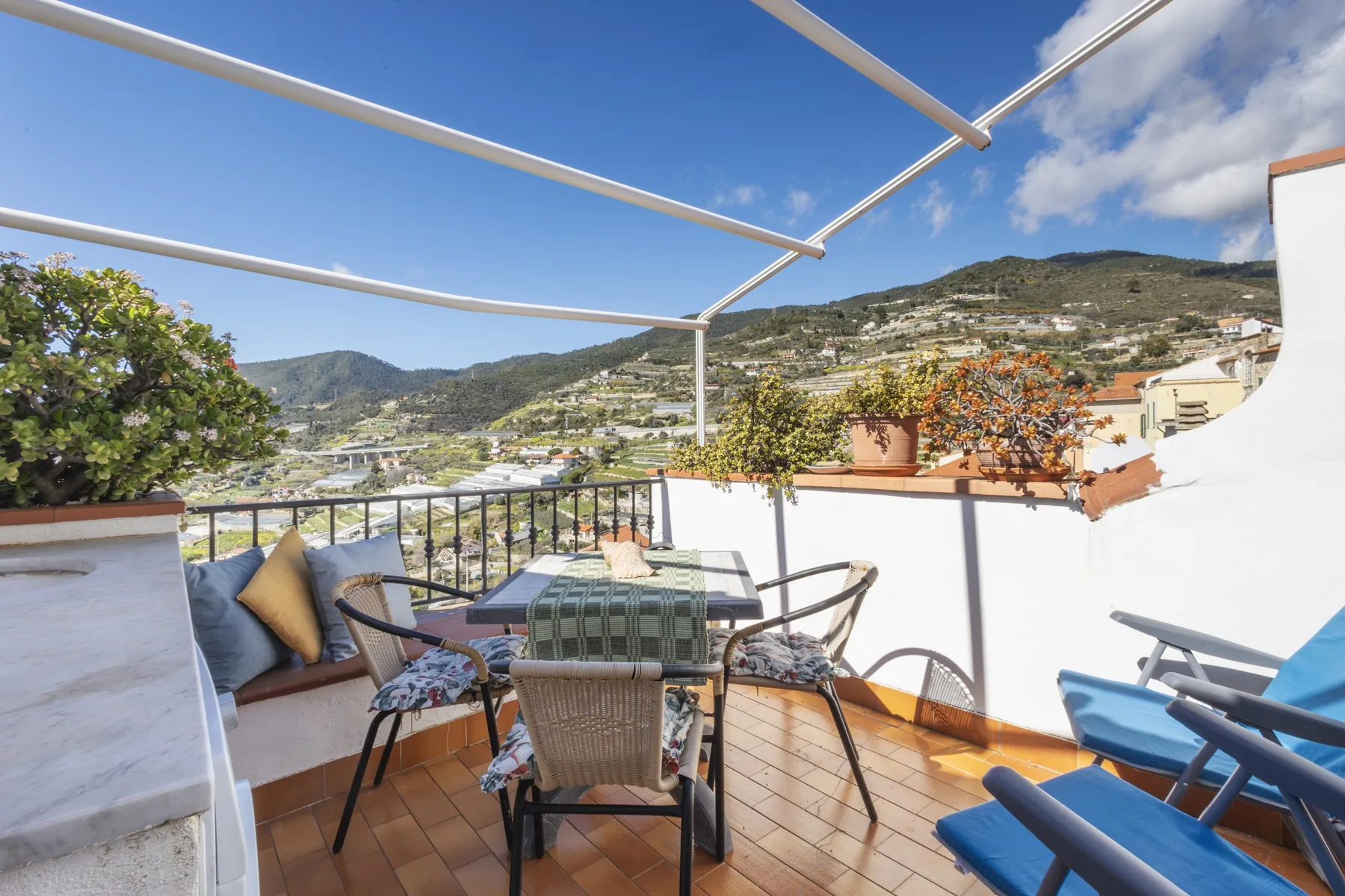 A tiled terrace with outdoor seating overlooks a hillside beneath a clear blue sky.