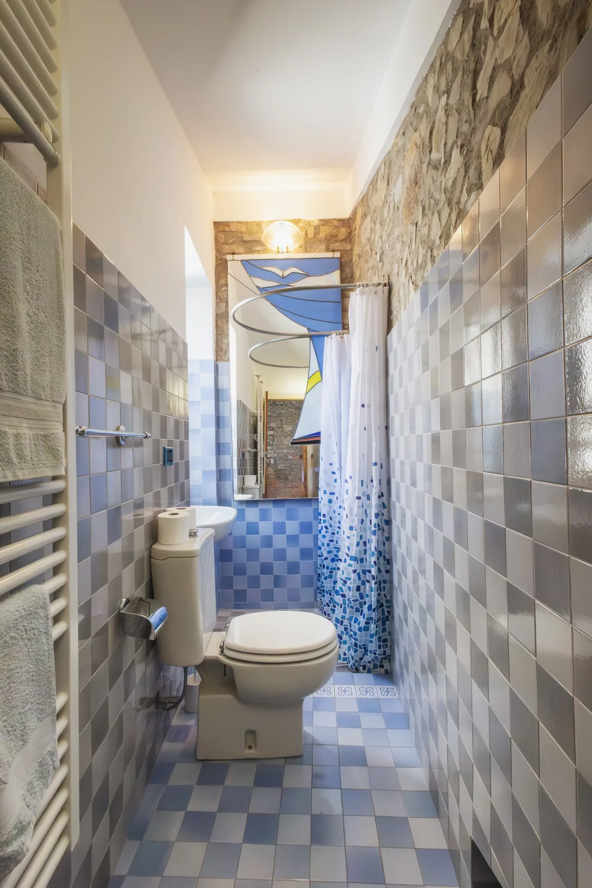A narrow tiled bathroom with a toilet, shower curtain, and wall-mounted towel warmer.
