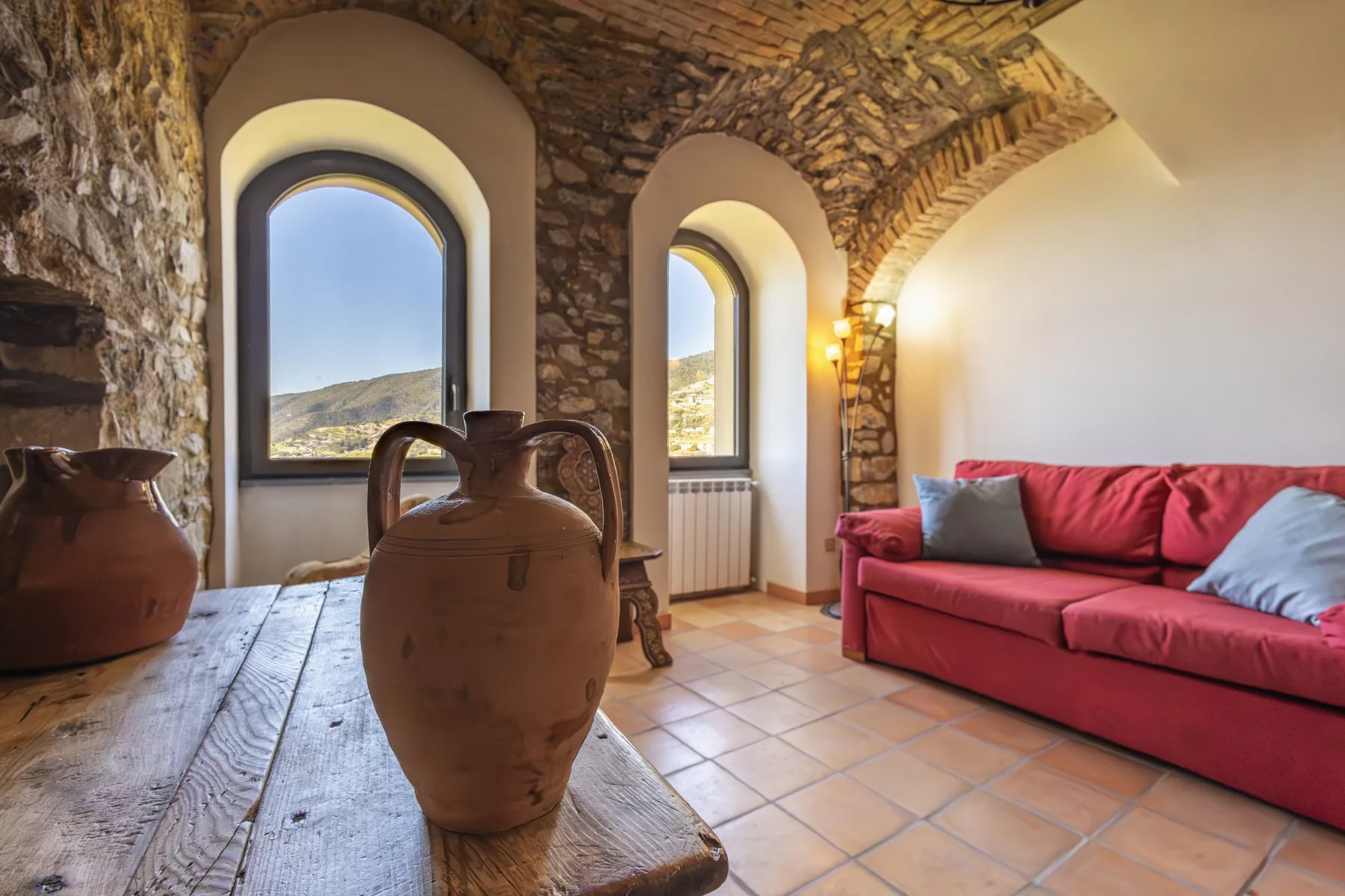 A rustic living room with a red sofa, arched windows, and stone walls.