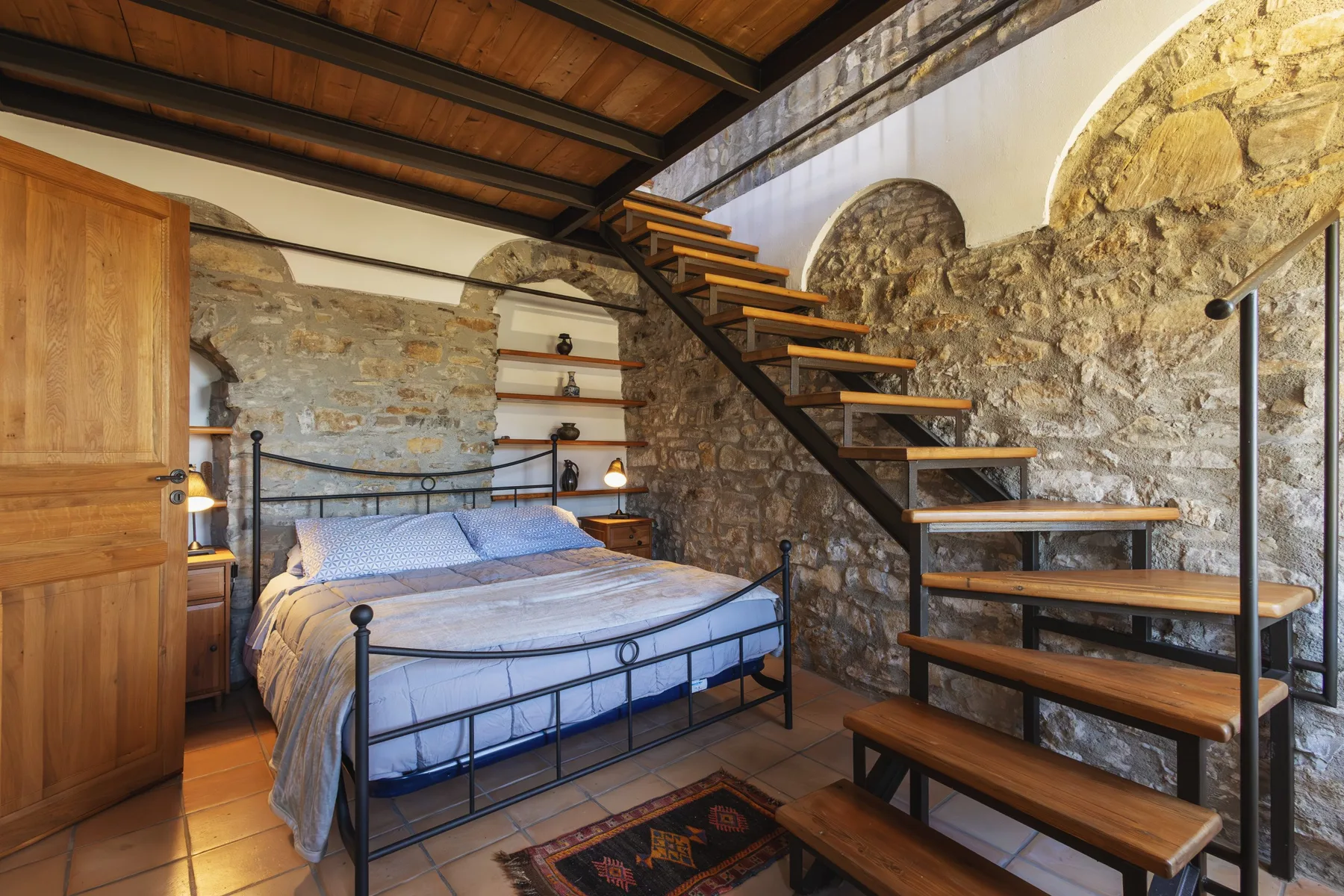 A bedroom with a bed beneath an open staircase and exposed stone walls.