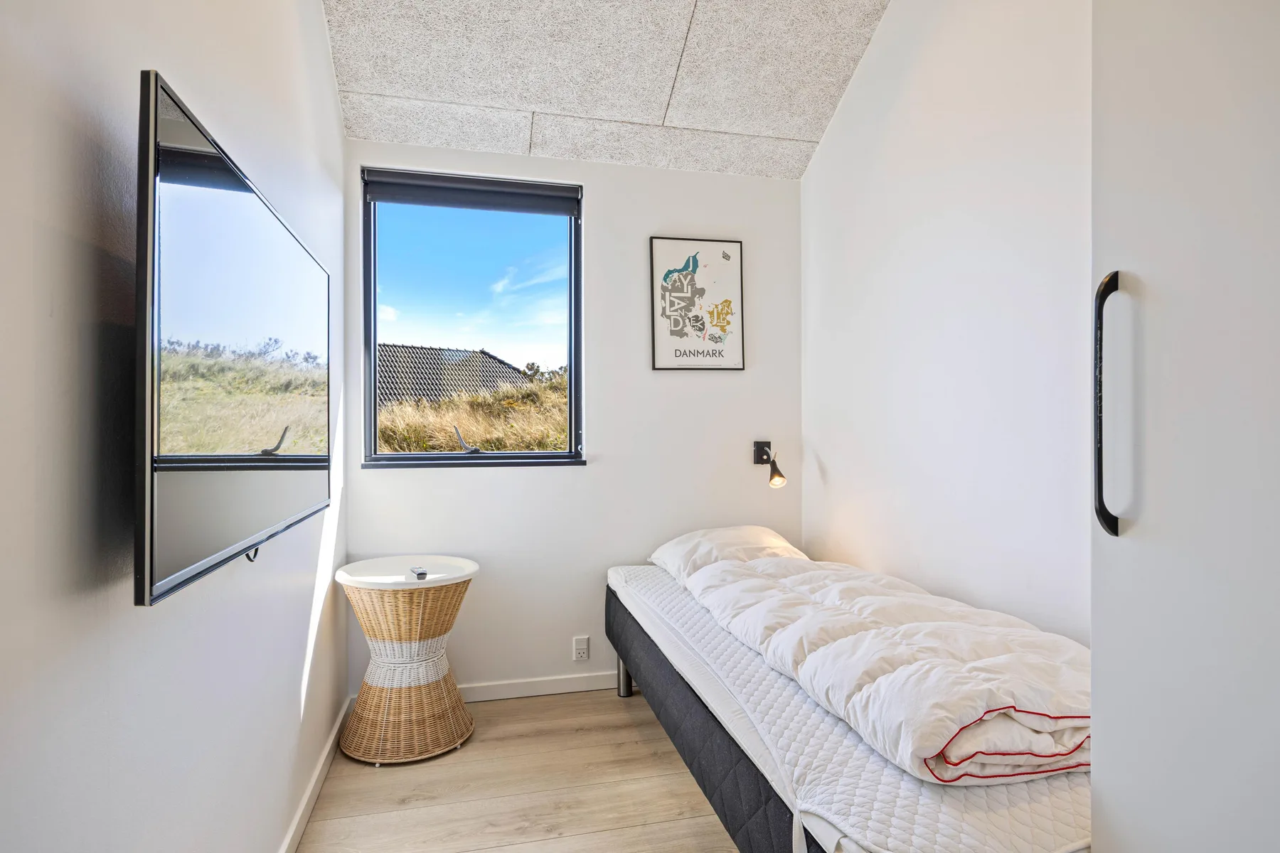 Un dormitorio luminoso con una cama individual, televisor en la pared y ventana.
