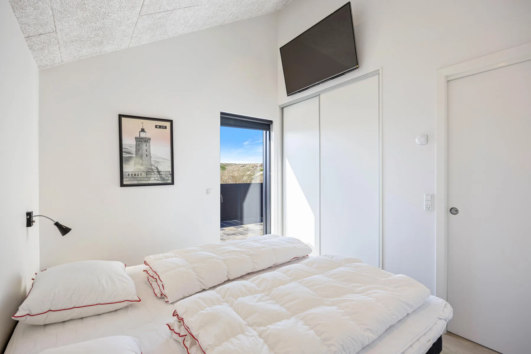 Un dormitorio luminoso con ropa de cama blanca y un televisor en la pared.