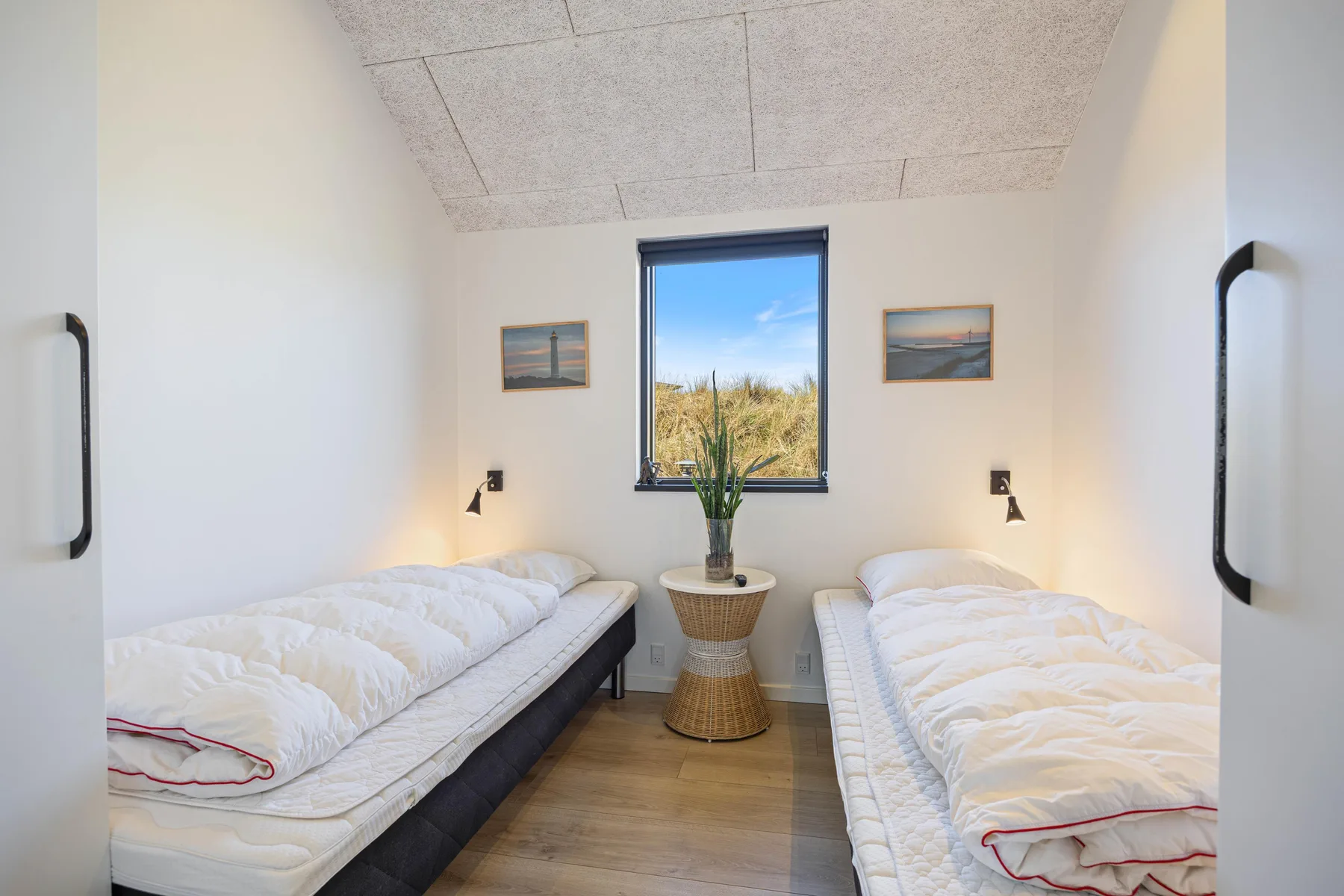 Un dormitorio luminoso con dos camas individuales y una ventana con vistas a dunas cubiertas de hierba.
