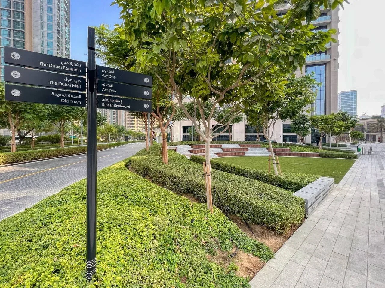 A landscaped pedestrian walkway with trees, hedges, and directional signs beside modern buildings.