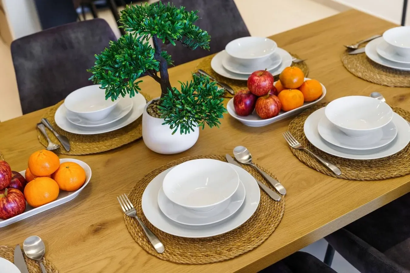 A dining table is set with white dishes, woven placemats, fruit, and a small potted plant.