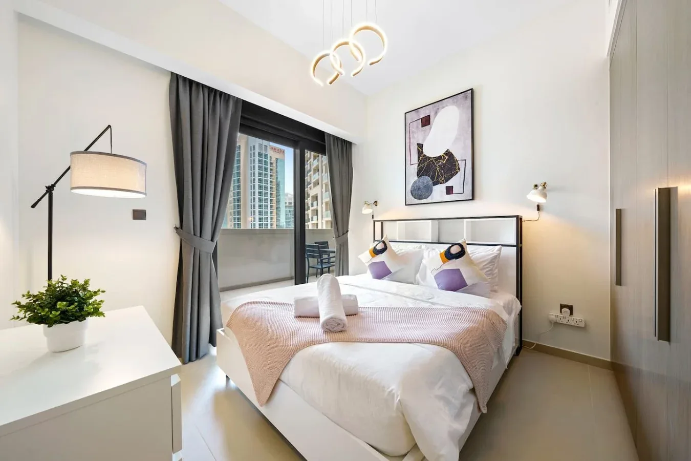 A bright bedroom with a neatly made bed, bedside lighting, and a window with curtains.