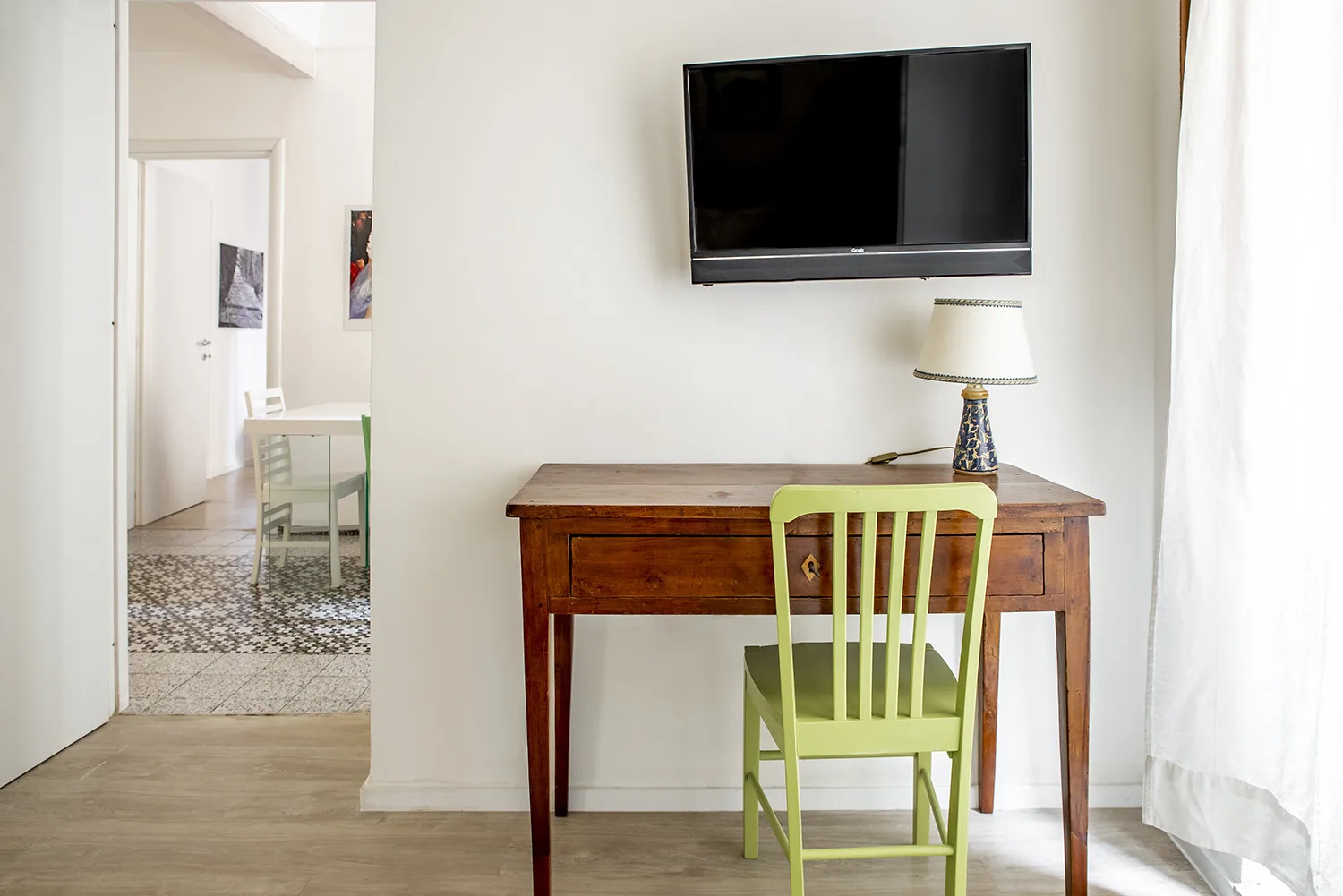 Un escritorio de madera con una silla verde y un televisor en la pared, en una habitación luminosa.