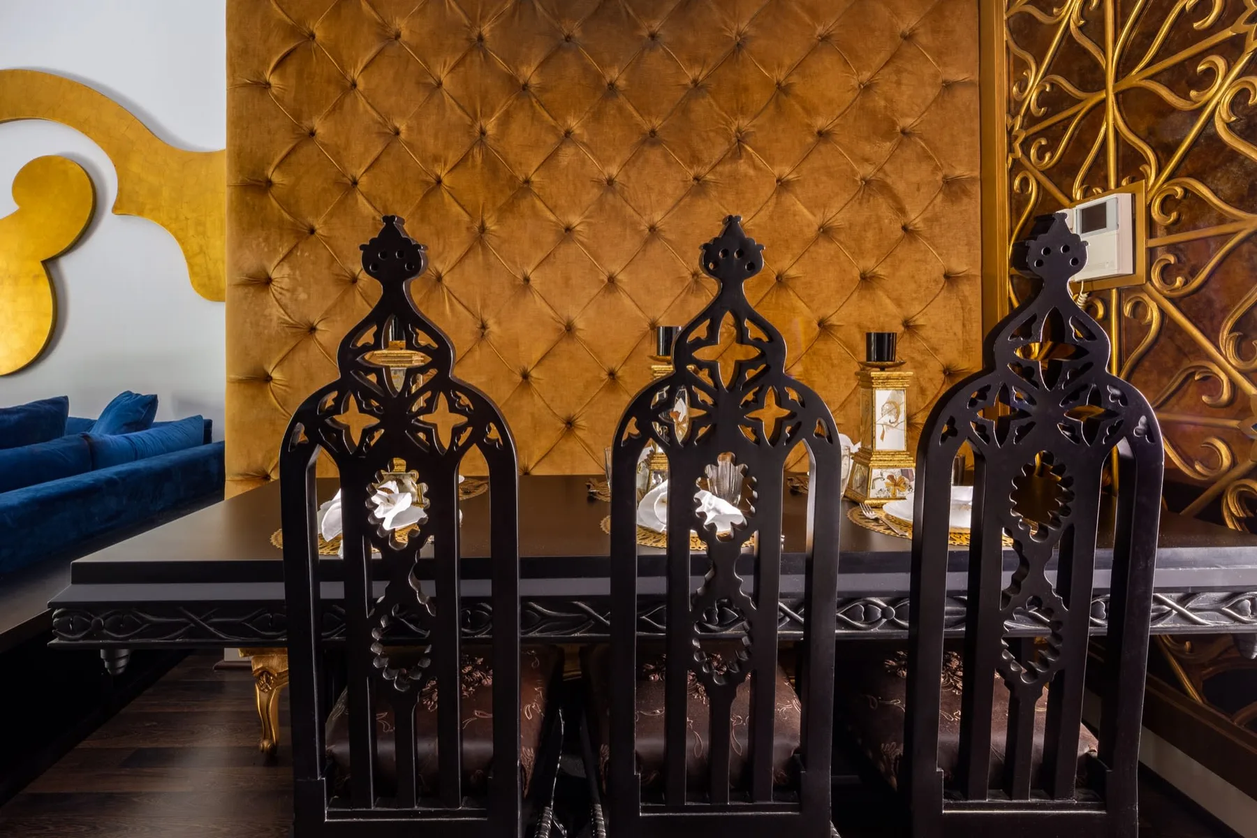 An ornate dining area with carved chairs in front of a tufted wall.