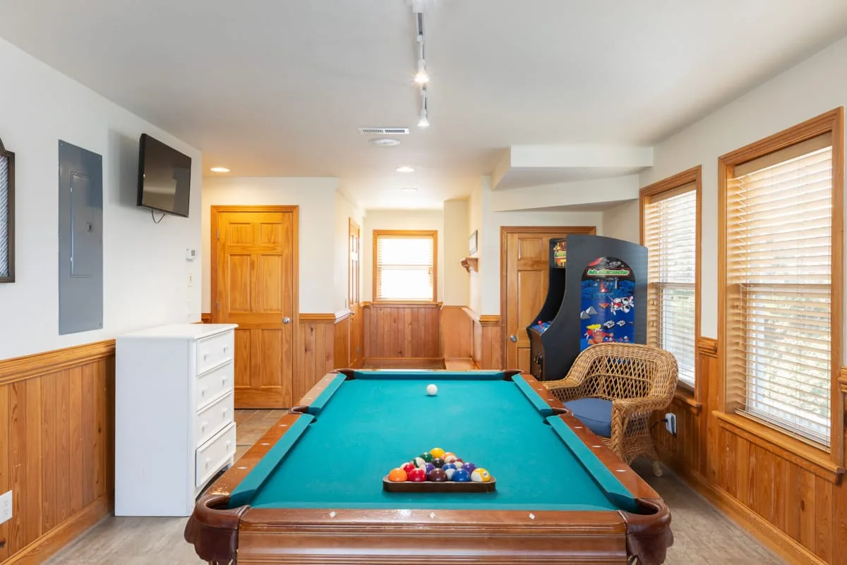 Una sala de juegos cuenta con una mesa de billar y una máquina recreativa junto a las ventanas.