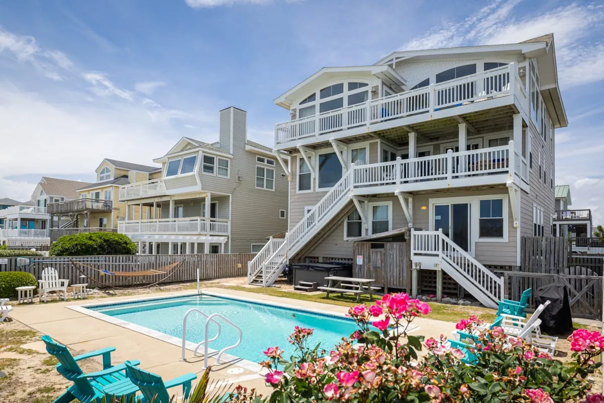 A beachside house overlooks a swimming pool, with outdoor seating and flowering plants.