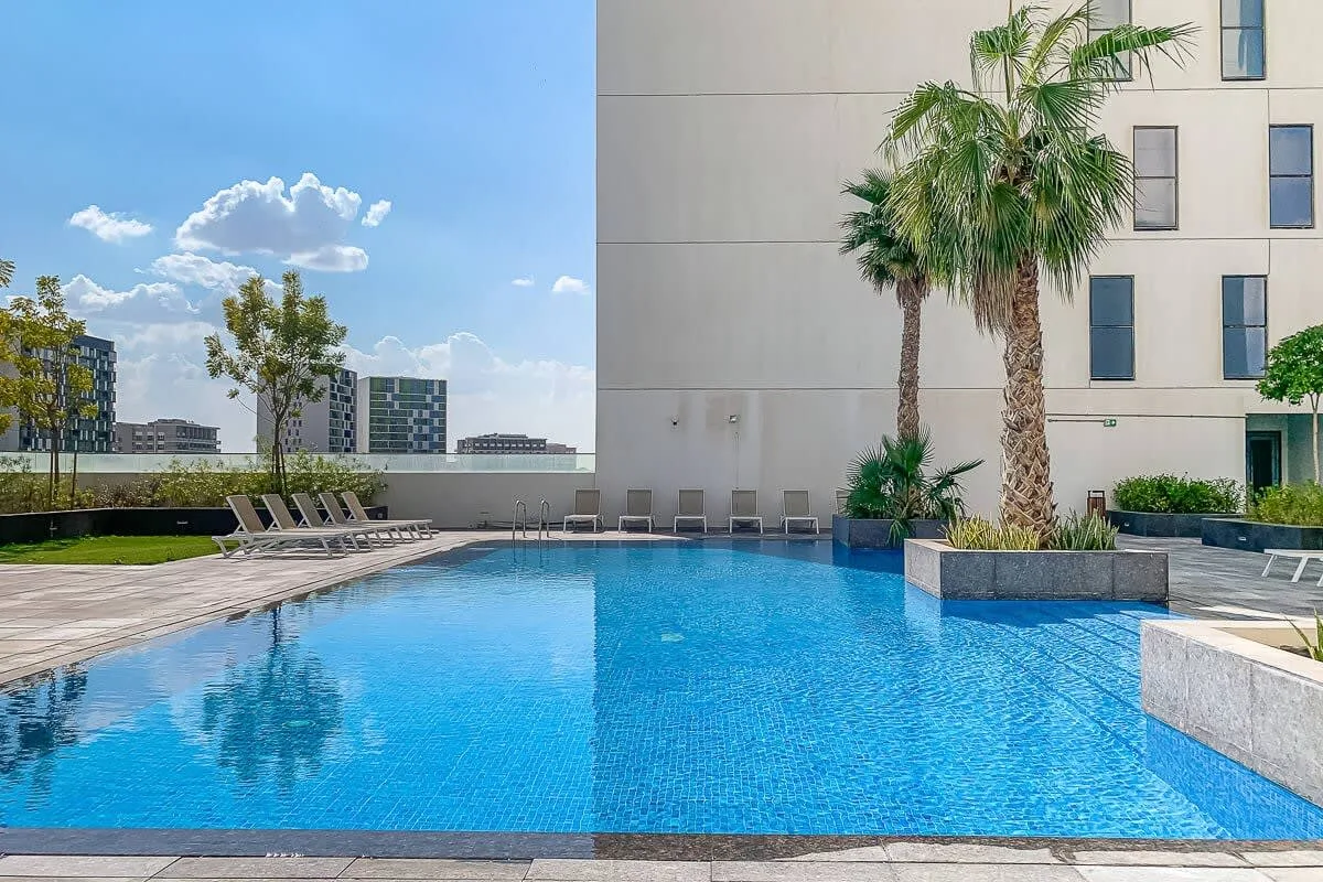 An outdoor swimming pool beside a modern building, with palm trees and lounge chairs.