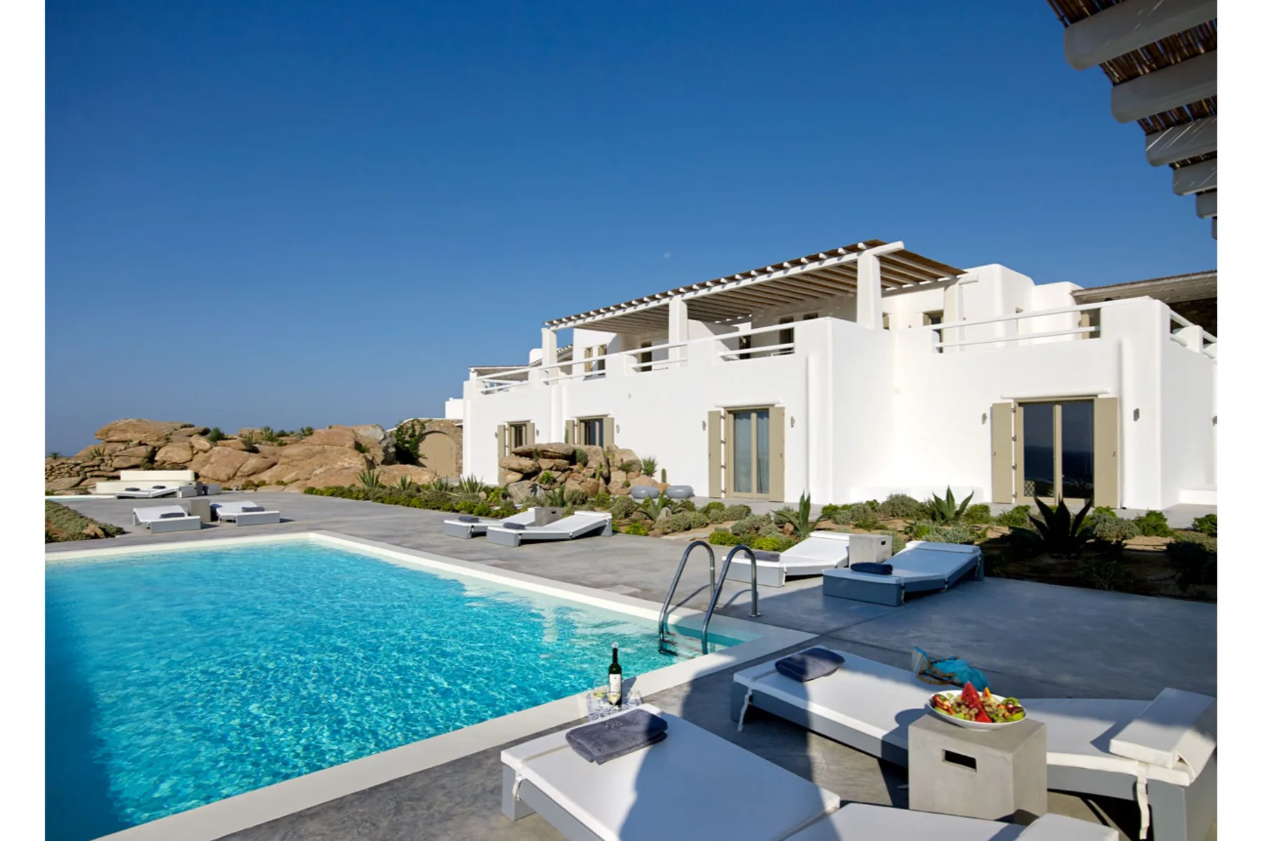 A white villa overlooks a swimming pool with sun loungers on the patio.
