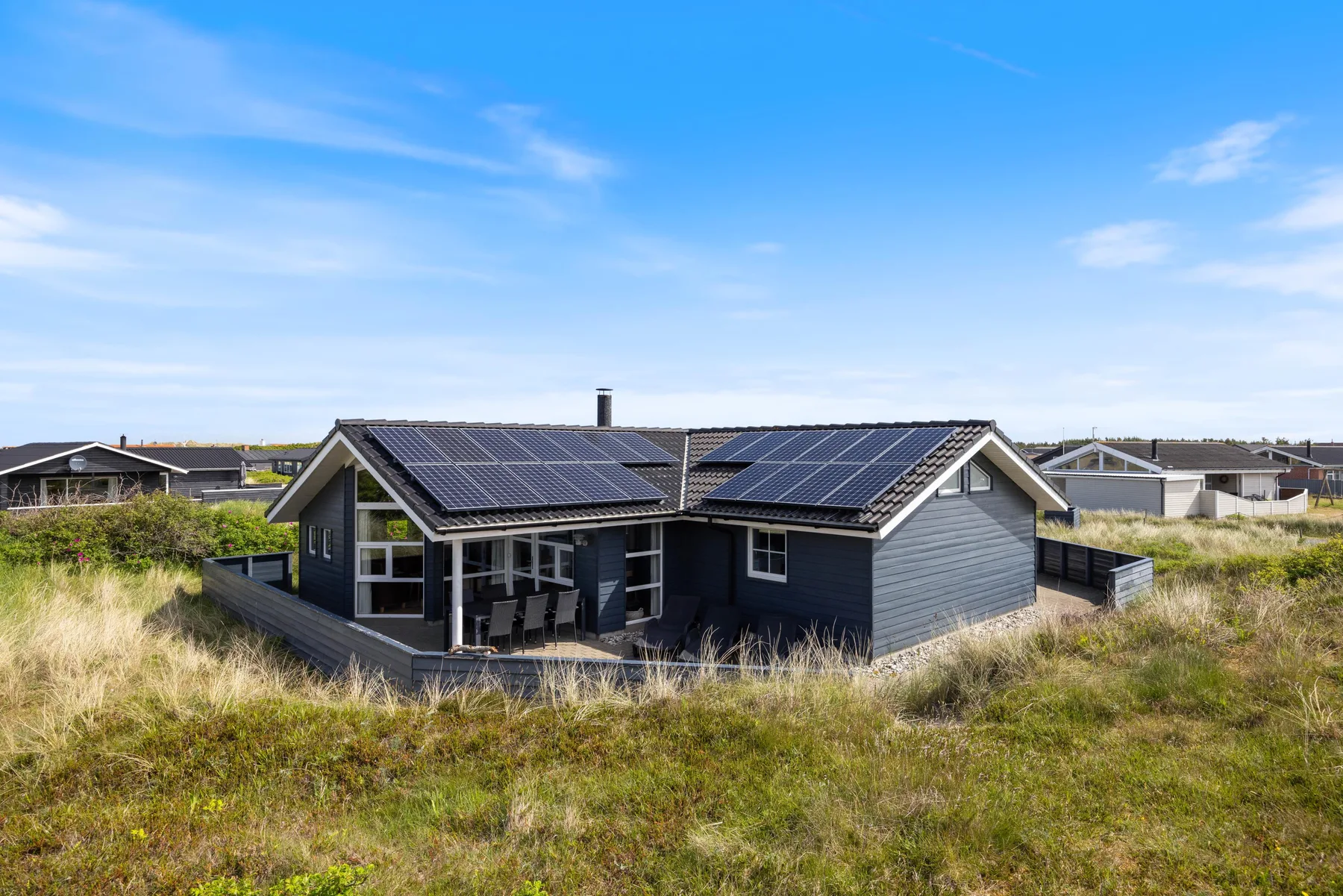 Una casa oscura con paneles solares se encuentra entre dunas cubiertas de hierba bajo un cielo azul.