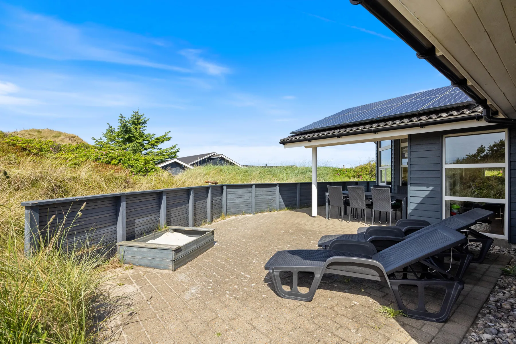 Un patio pavimentado junto a una casa tiene asientos al aire libre y una jardinera baja de madera.