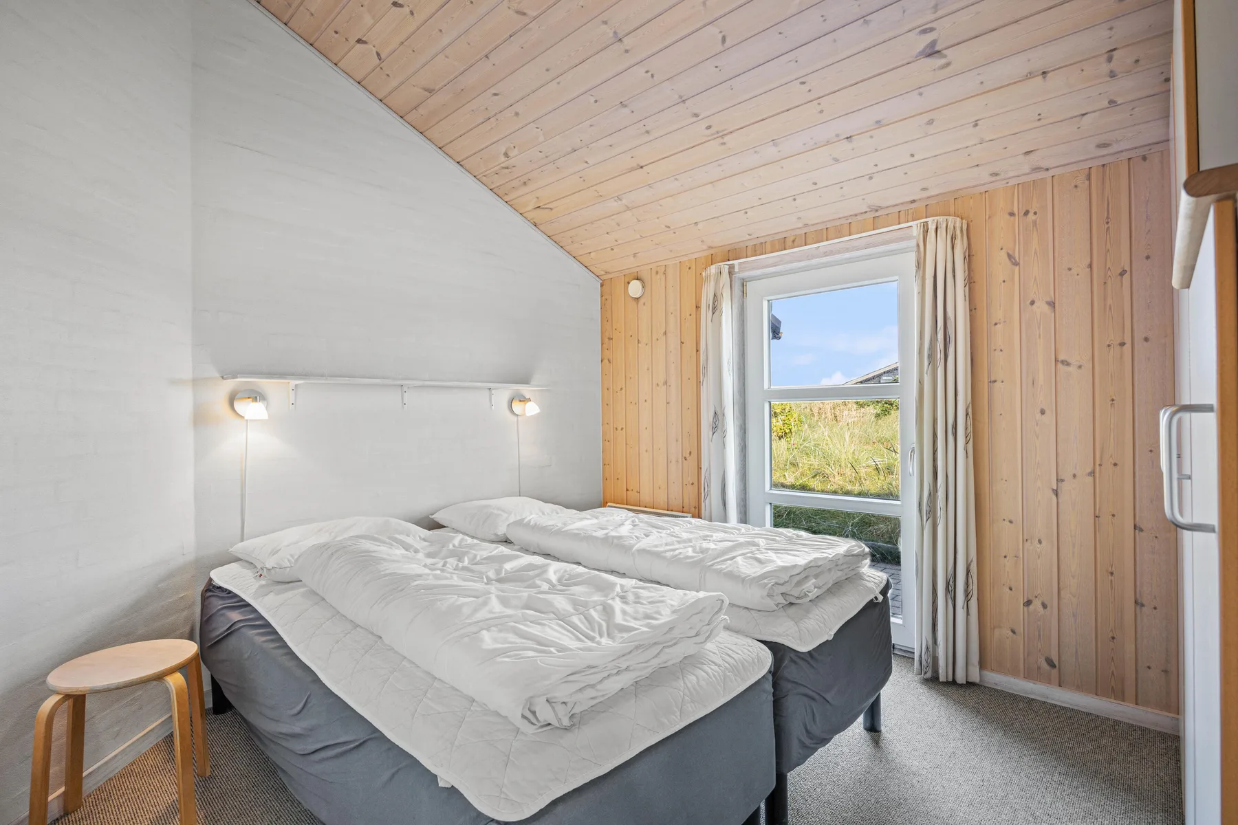 Un dormitorio con dos camas, luces junto a ellas y una ventana con vistas a la vegetación.