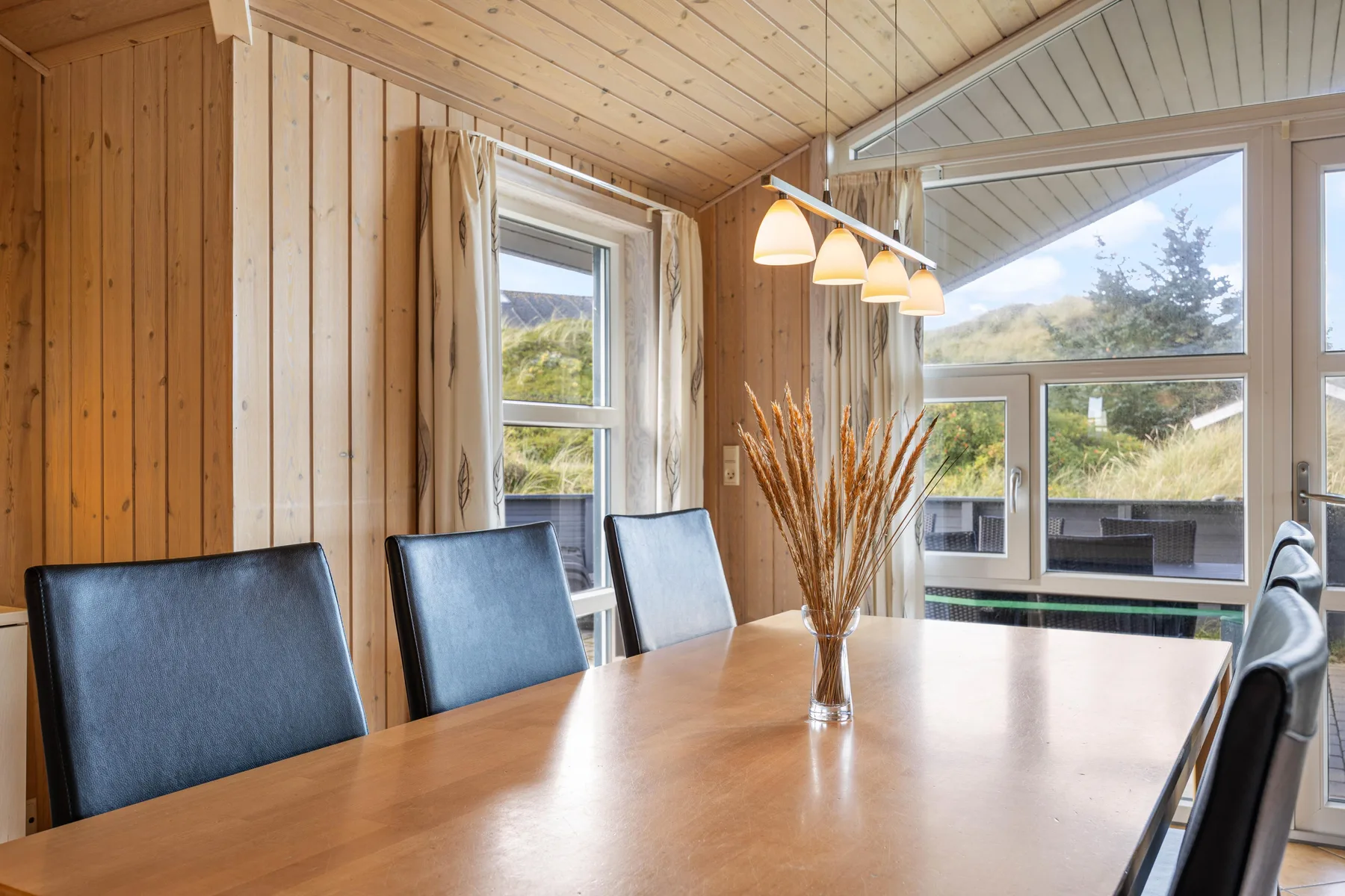 Una zona de comedor tiene una mesa larga de madera junto a grandes ventanas.
