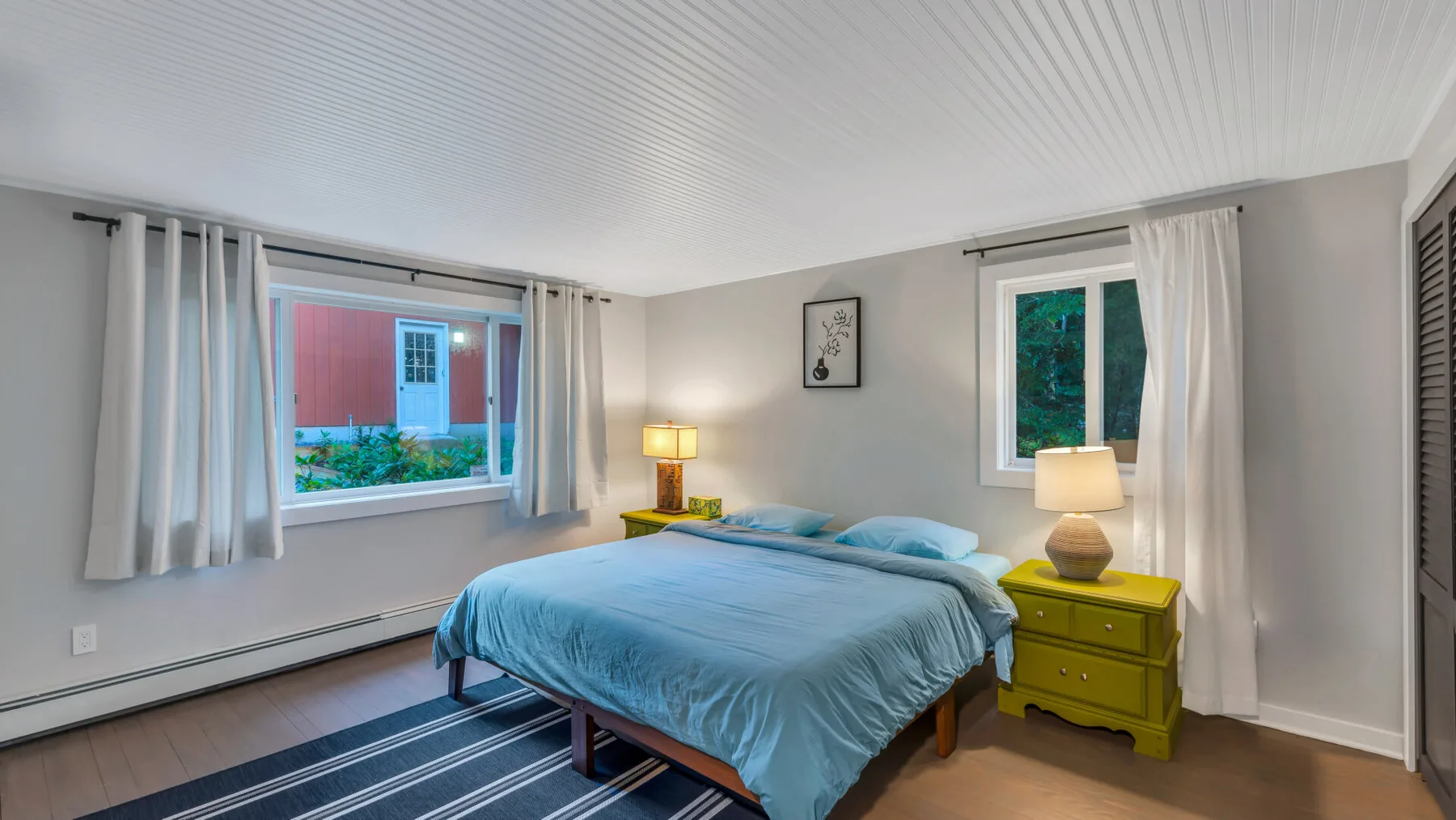 A bedroom with a bed between two curtained windows and a bedside lamp.