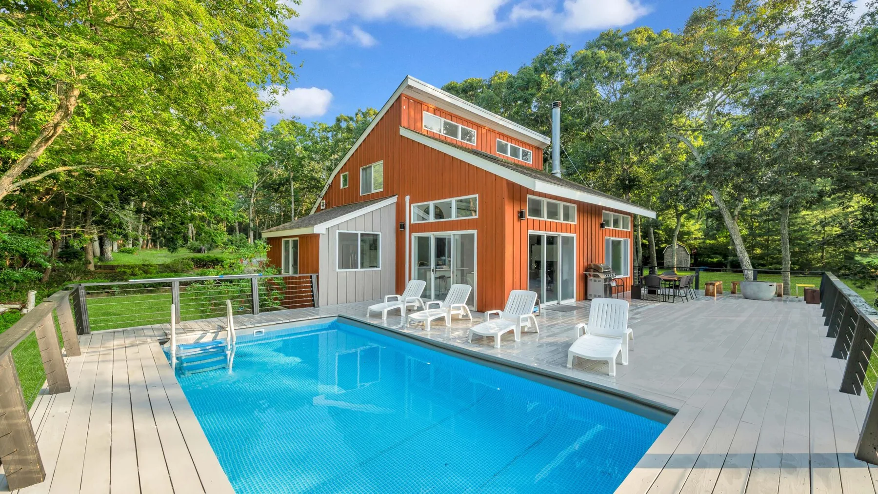 A red house stands beside a swimming pool, with trees surrounding the outdoor patio.