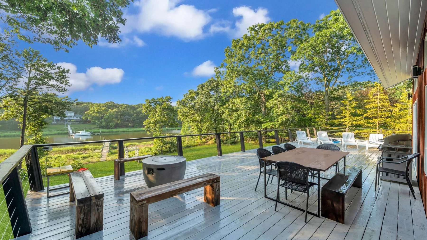 A lakeside deck has a dining table, seating, and a hot tub.