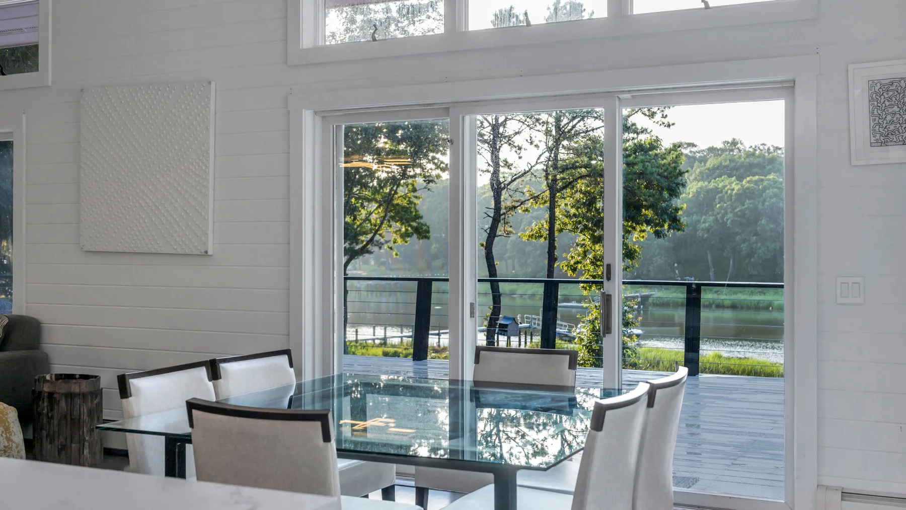 A bright dining area with a glass table beside large windows overlooking greenery.