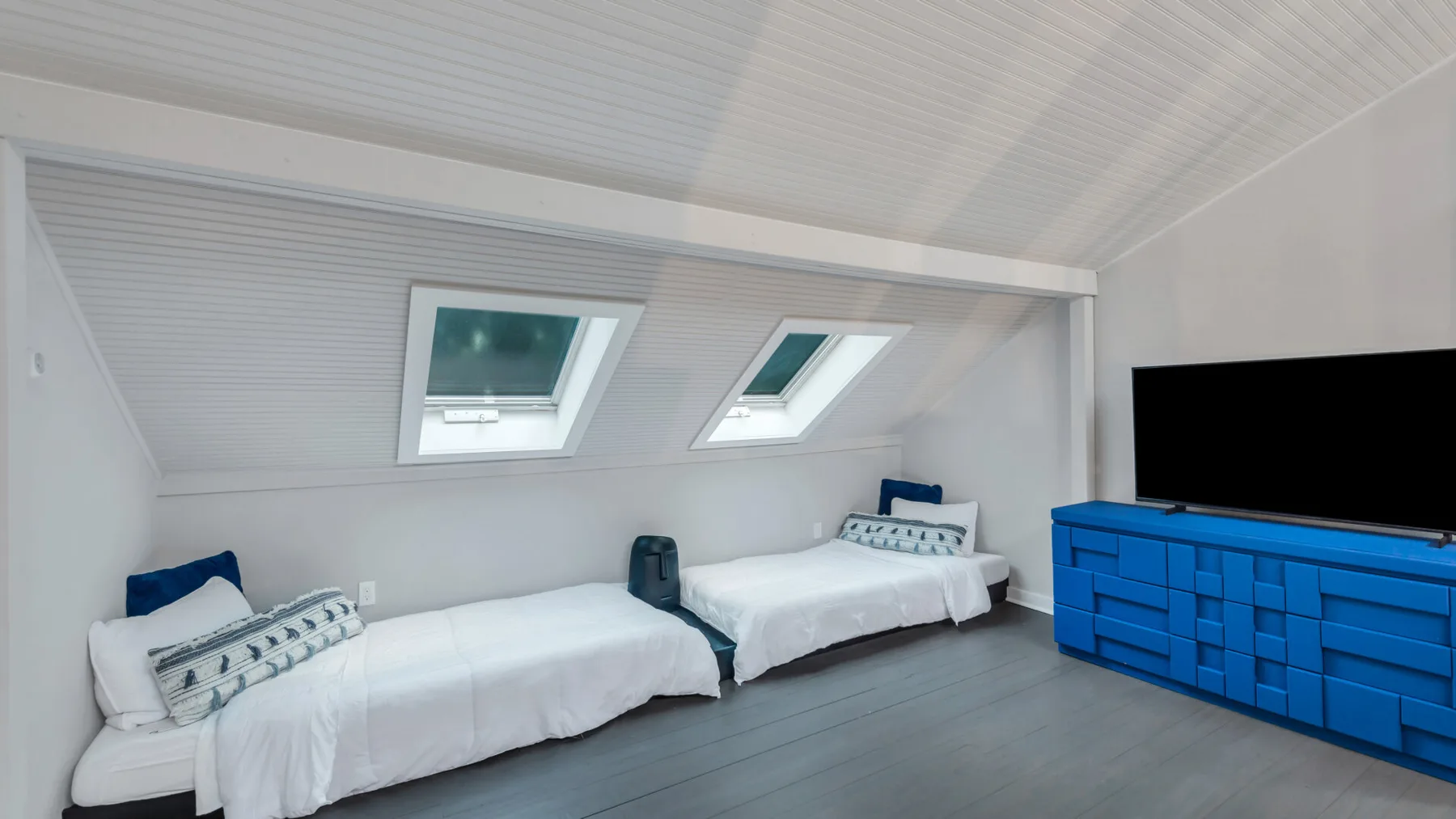 A bright bedroom with two beds beneath sloped ceilings and a blue dresser.