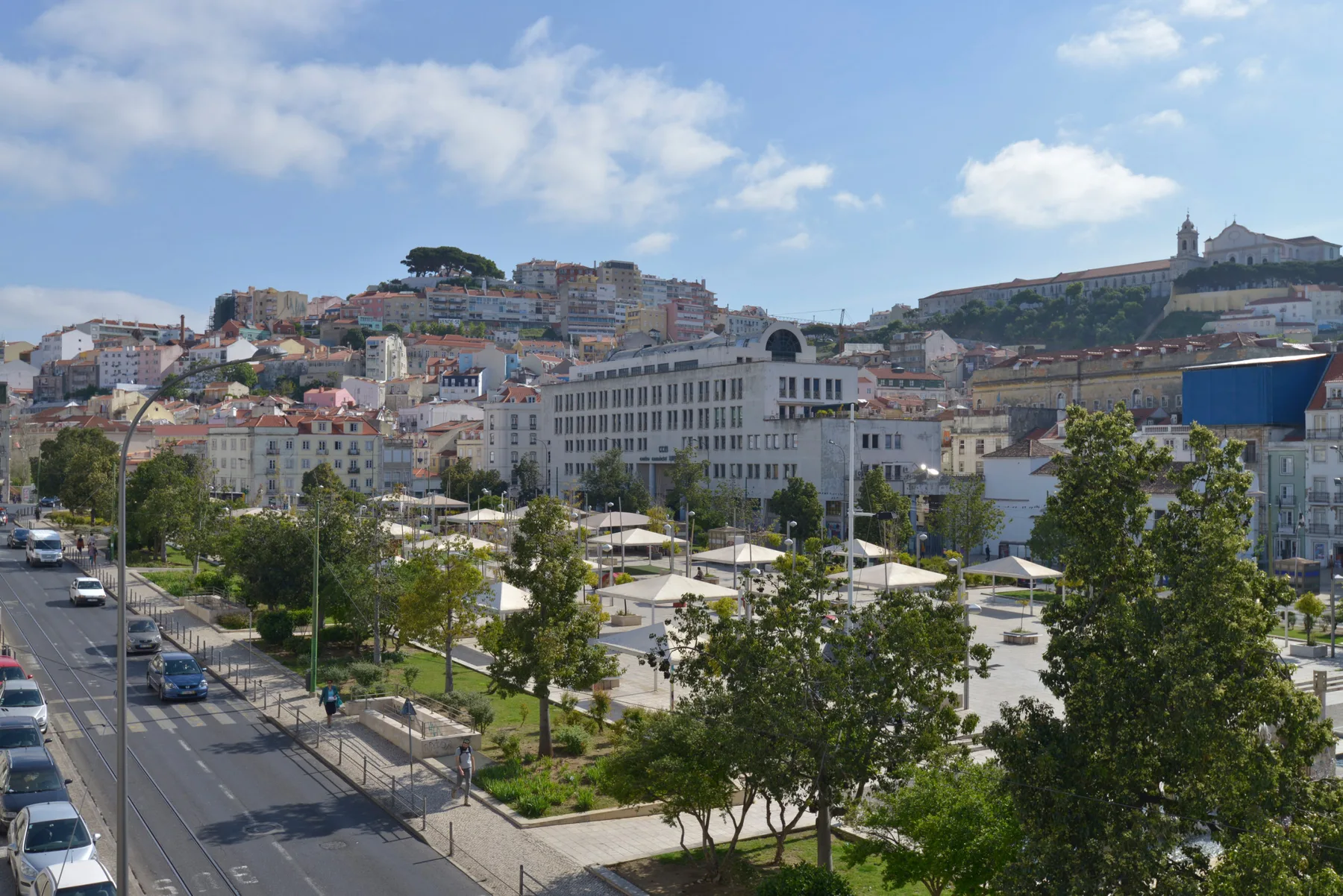 Una plaza urbana con árboles y asientos, rodeada de edificios en una ladera y un castillo en lo alto.