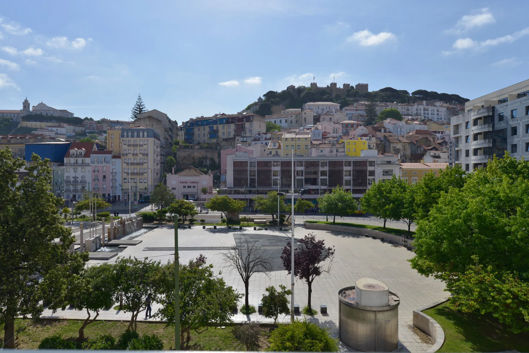 Una plaza urbana con árboles y edificios, con un castillo en lo alto de una colina al fondo.
